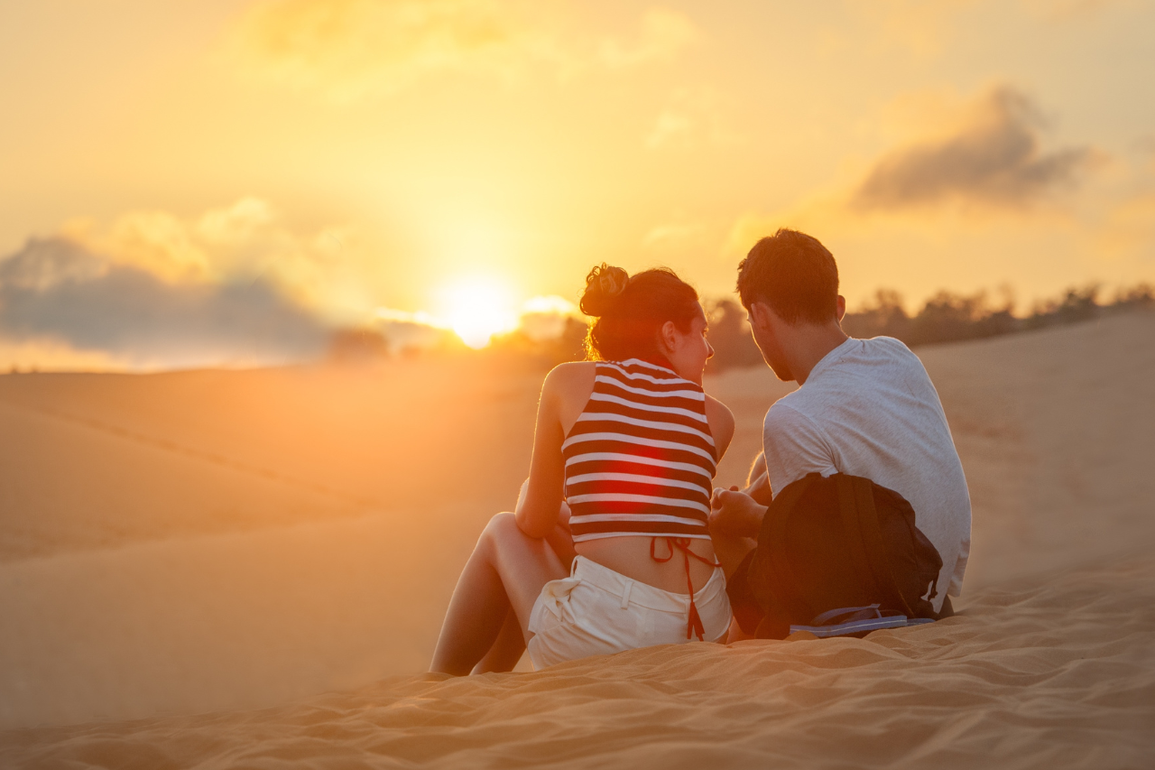 Man & woman sitting on a sand dune watching the sun set. 